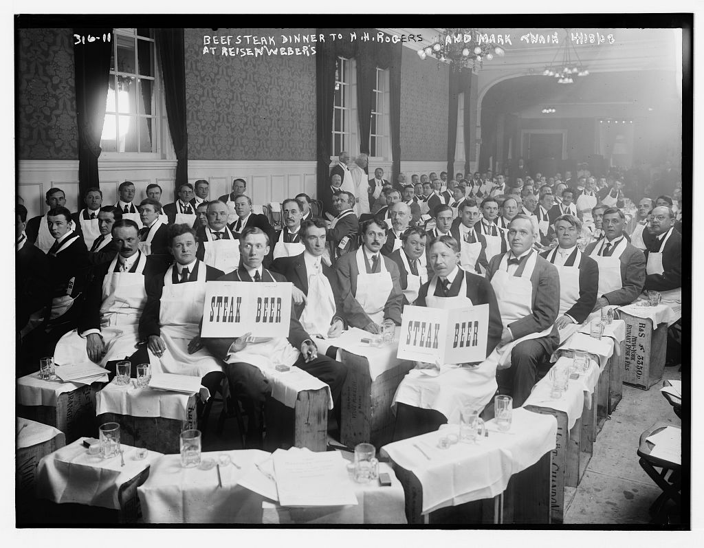 Guests in white aprons hold STEAK and BEER signs at a 1908 Beefsteak dinner at Reisenweber’s in New York